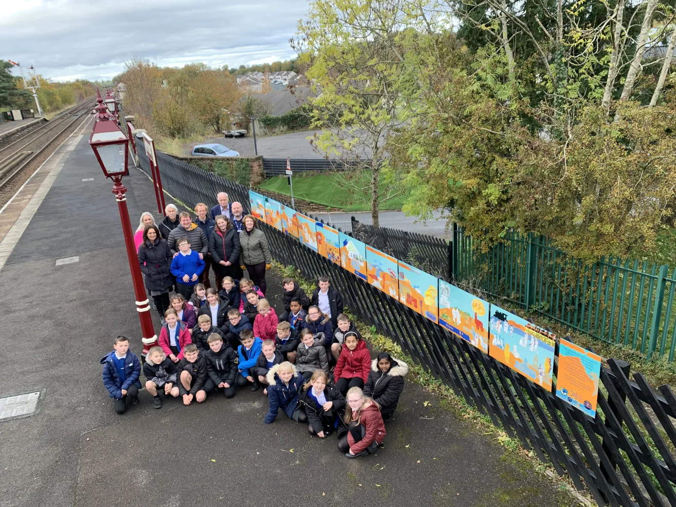A large group of children on a railway platform with a large piece of artwork at Appleby Station. Appleby Primary scvhool with artist Karen Babayan
