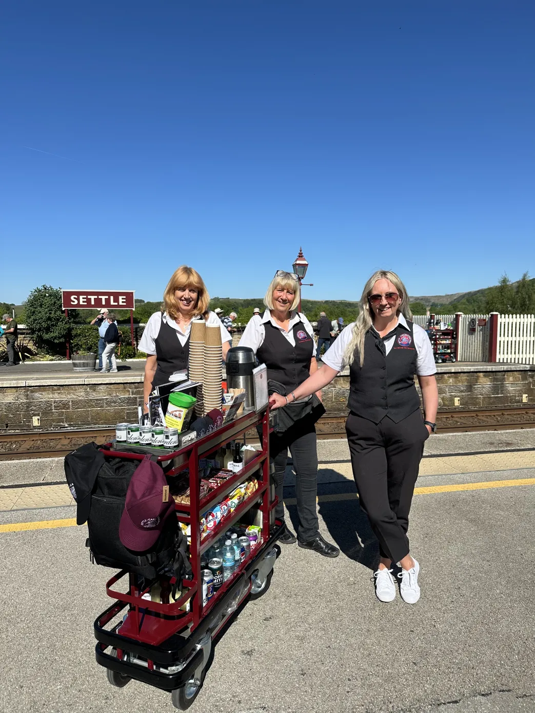 Blue sky and sign saying Settle, with three women next to a refreshment trolley.