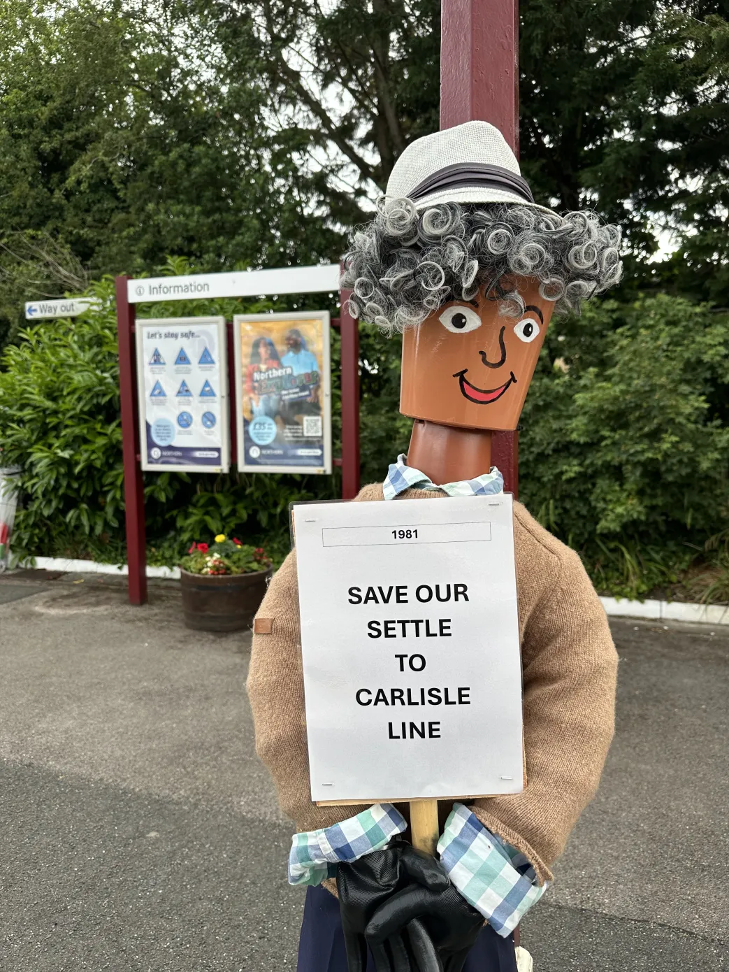 Character made of flowerpots on a station platform with a 'Save our Settle to Carlisle Line' poster