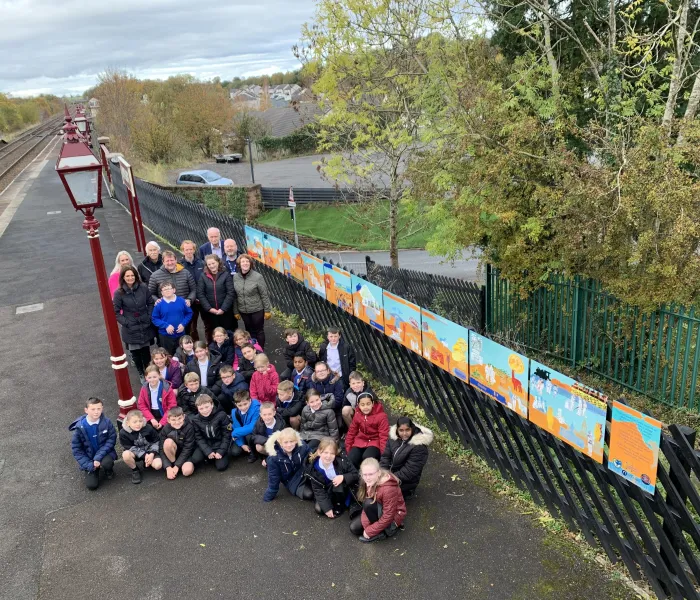 A large group of children on a railway platform with a large piece of artwork at Appleby Station. Appleby Primary scvhool with artist Karen Babayan
