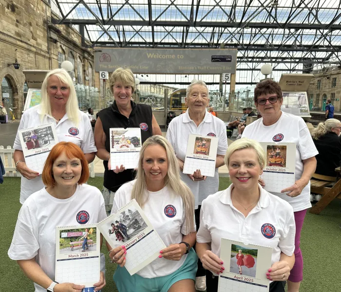 Seven women in martching t-shirt holding copies of a calendar in Carlisle Railway Station