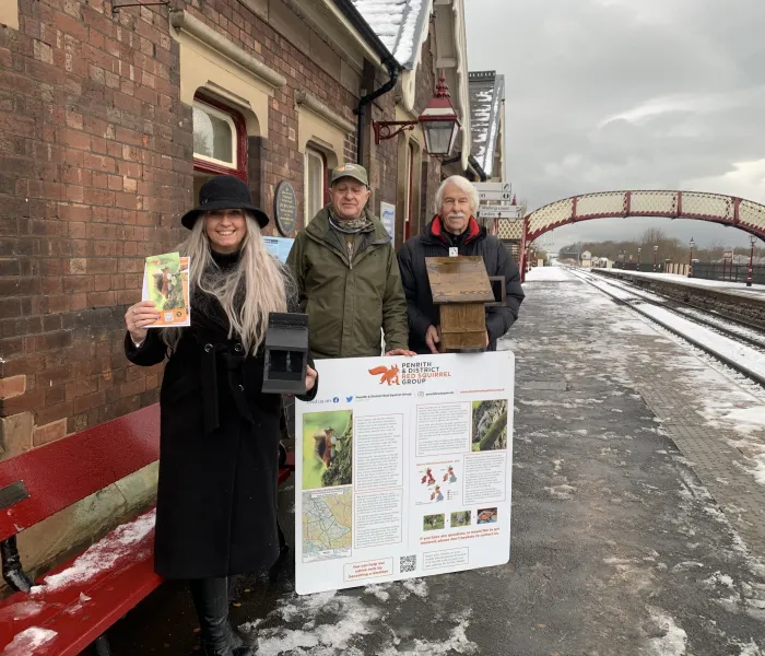 a woman and 2 men on a railway station platform with an onformation board about red squirrels
