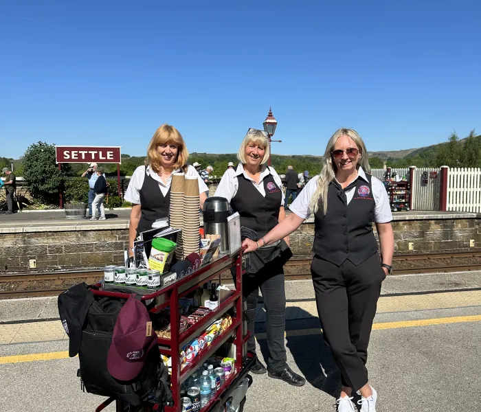 Blue sky and sign saying Settle, with three women next to a refreshment trolley.
