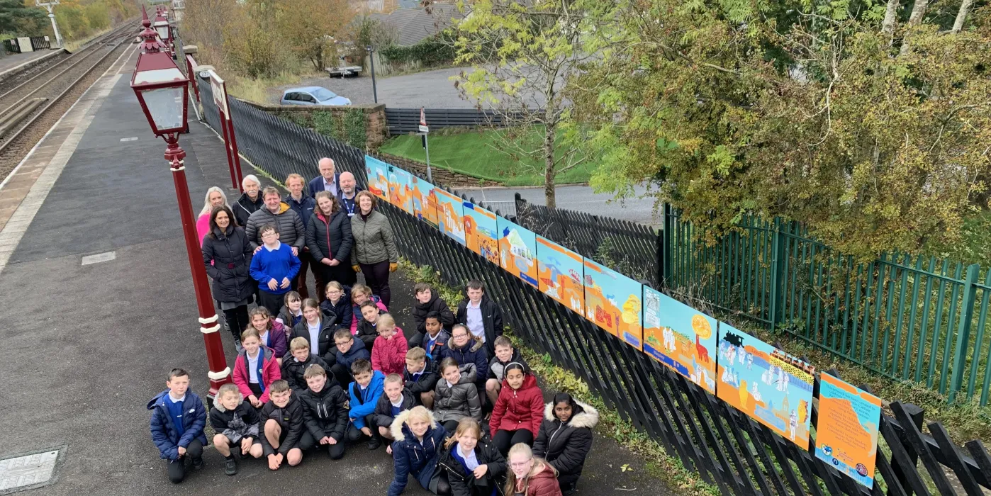 A large group of children on a railway platform with a large piece of artwork at Appleby Station. Appleby Primary scvhool with artist Karen Babayan