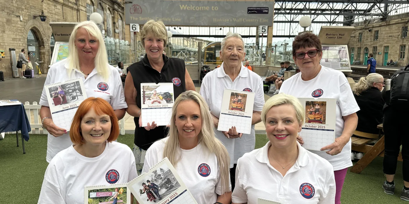 Seven women in martching t-shirt holding copies of a calendar in Carlisle Railway Station