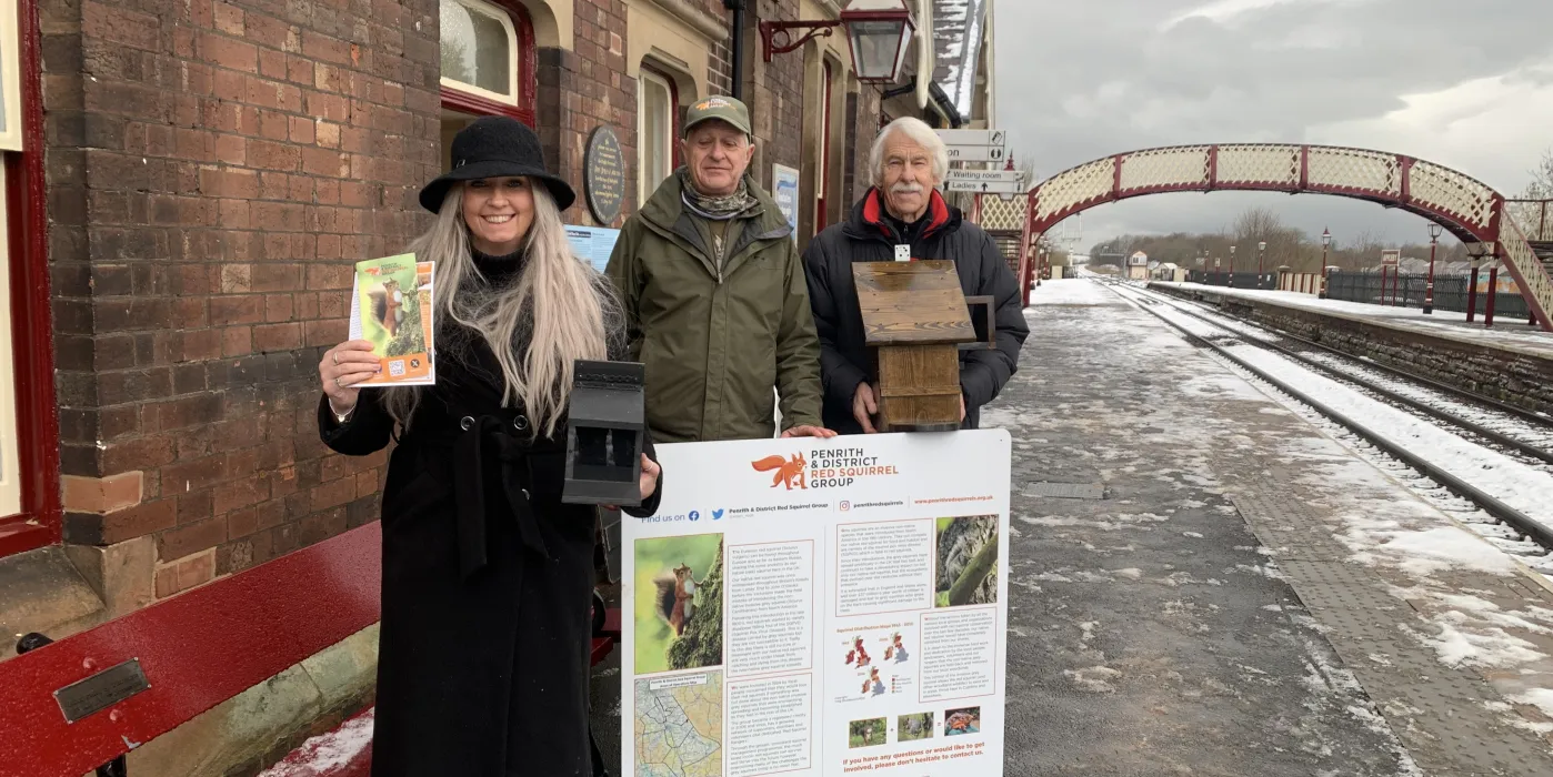 a woman and 2 men on a railway station platform with an onformation board about red squirrels