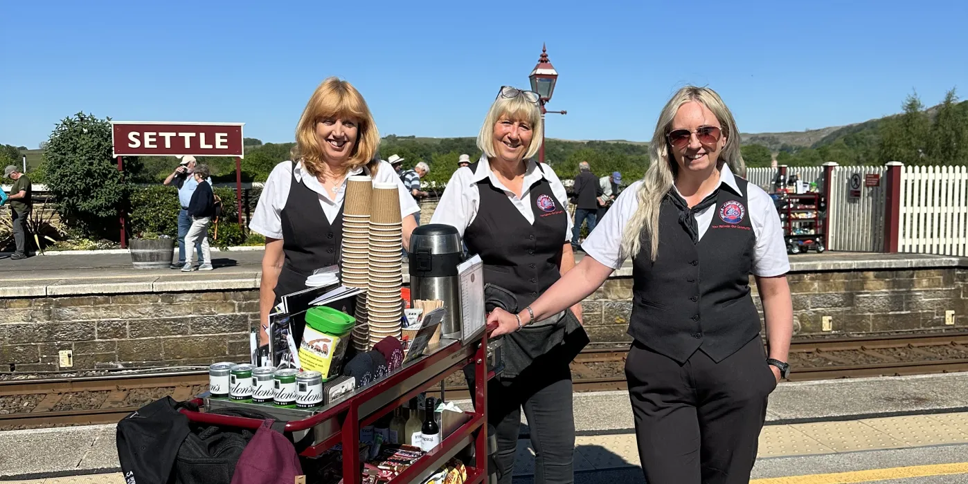 Blue sky and sign saying Settle, with three women next to a refreshment trolley.