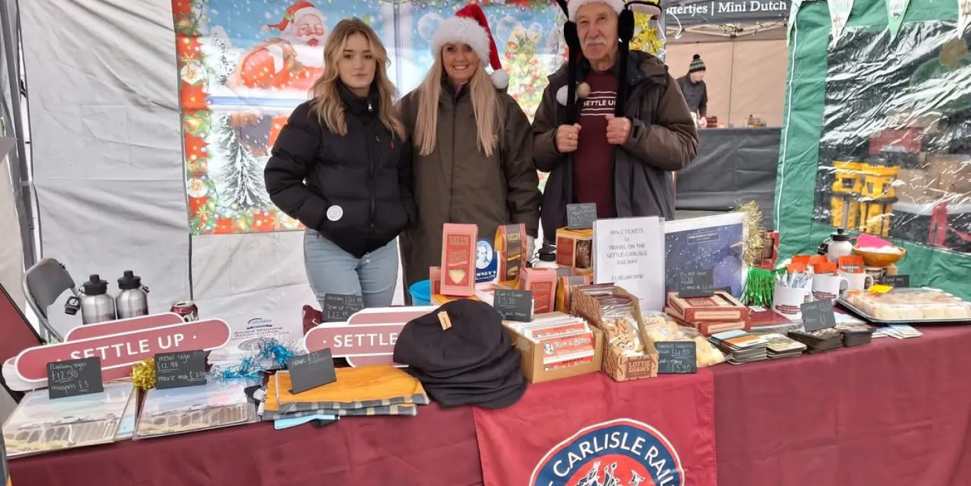 Three people, with santa hats on a stall with Settle Carlisle Railway logo